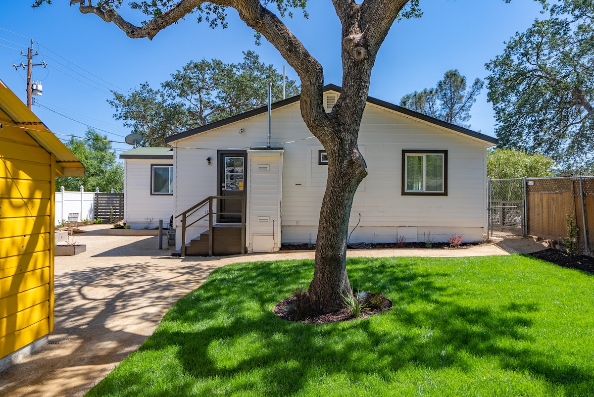 The exterior of the home is shown, featuring a well-maintained lawn and a decorative tree. A pathway leads to the entrance, with a light-colored structure visible. A fenced area is present in the background, creating a sense of privacy.