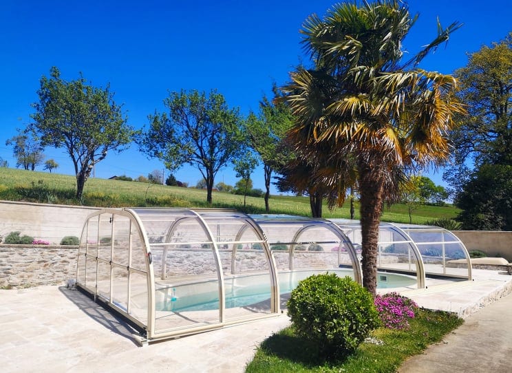 A private pool is enclosed within a transparent cover, surrounded by landscaped greenery. Palm trees and flowering shrubs add a natural touch to the outdoor space. A clear blue sky enhances the tranquil setting, promoting a serene atmosphere for relaxation.