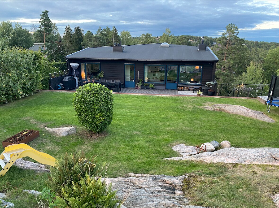 A modern black house is set against a backdrop of greenery, surrounded by a well-maintained garden featuring a variety of plants and grassy areas. Natural rock formations and a slide are visible in the foreground, with a patio and outdoor seating area situated in front of the large windows.