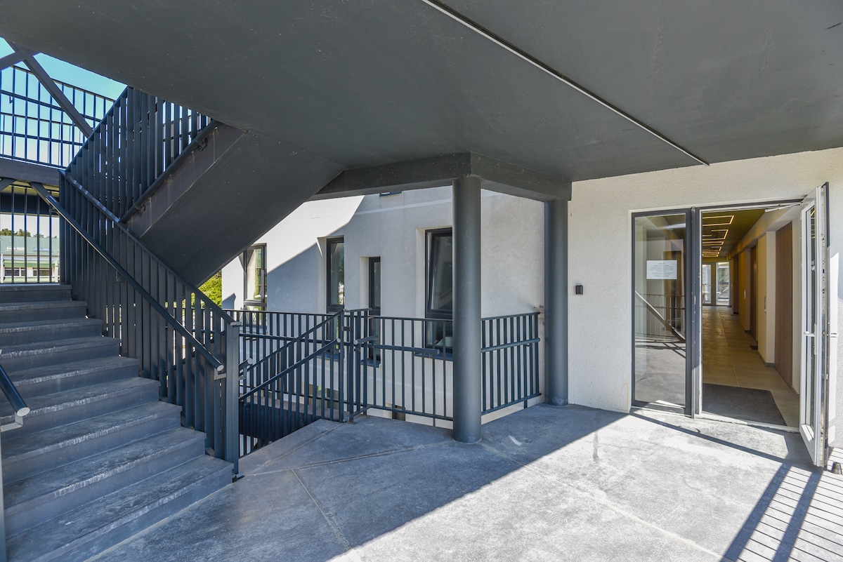 An exterior view of a multi-level building entrance is displayed, featuring a covered patio area. A set of stairs is positioned on the left, leading to the upper floors. Double doors provide access into the building's interior, framed by a long, open walkway.
