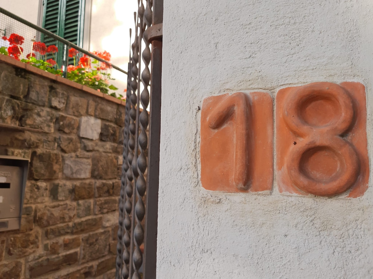 A close-up of the building number '18' is displayed in terracotta on a white wall. A wrought iron gate is visible, with a glimpse of a stone wall and vibrant red flowers in the background, adding a touch of color to the setting.