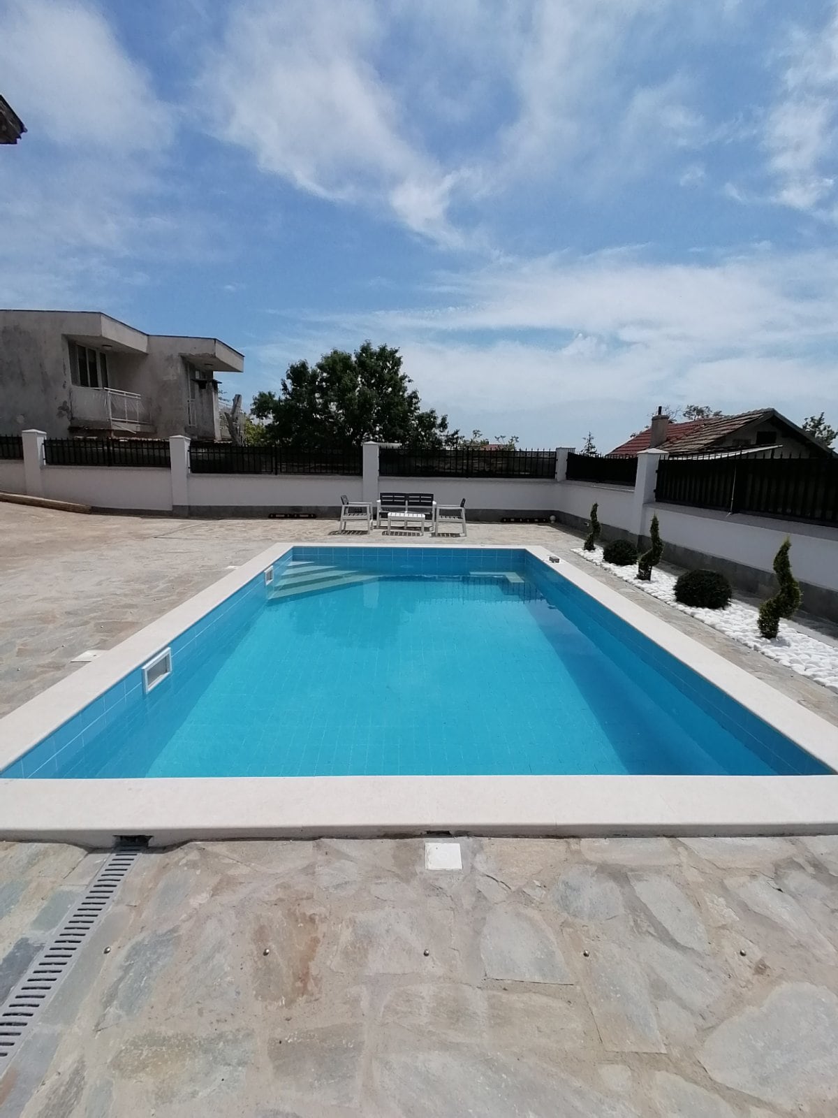 A rectangular outdoor swimming pool is surrounded by a paved area and low landscaping. Several lounge chairs are arranged on one side of the pool, and a clear blue sky is visible above, creating a bright and open atmosphere.