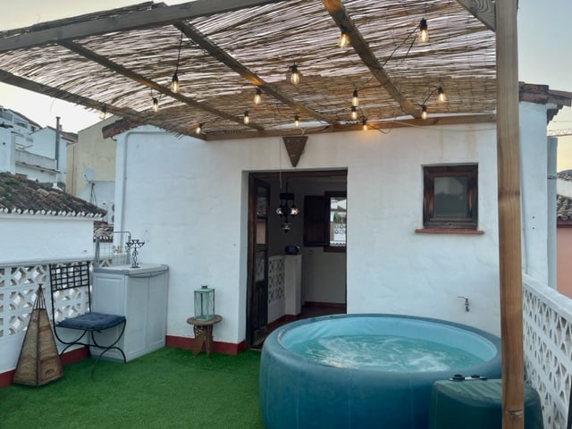 A spacious roof terrace is fitted with a Jacuzzi, surrounded by greenery and lighted with string lights. A shaded area is created by a bamboo structure, and a doorway opens into the interior of the townhouse. Outdoor seating is also present.