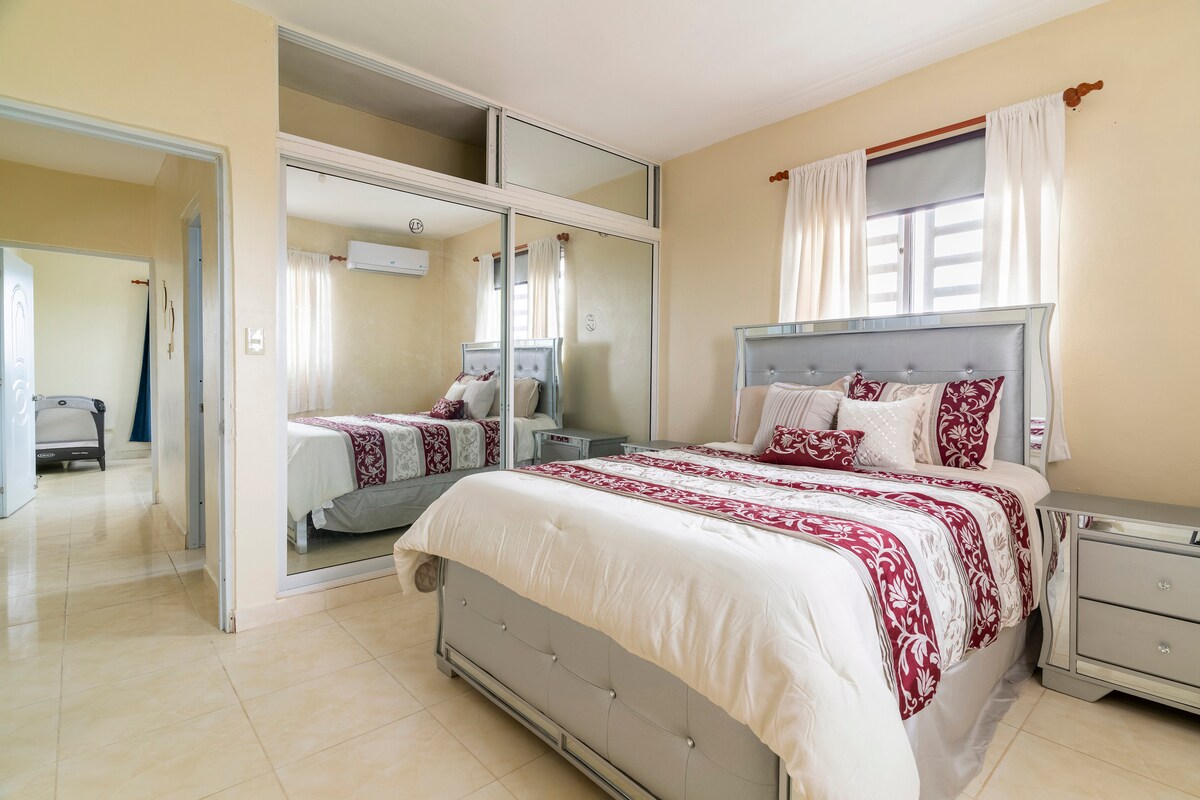 A spacious bedroom features a plush bed dressed in a decorative red and white coverlet. Mirrored closet doors reflect the light from large windows, enhancing the bright and airy feel of the room. Soft beige walls complement the modern furnishings.