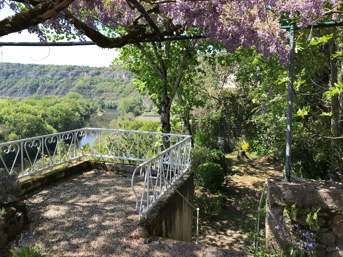 A gravel terrace is framed by lush greenery and flowering plants, offering views of the river below. Wisteria drapes elegantly from the overhead structure, enhancing the tranquil outdoor space. A winding pathway leads through the garden, inviting exploration of the natural surroundings.