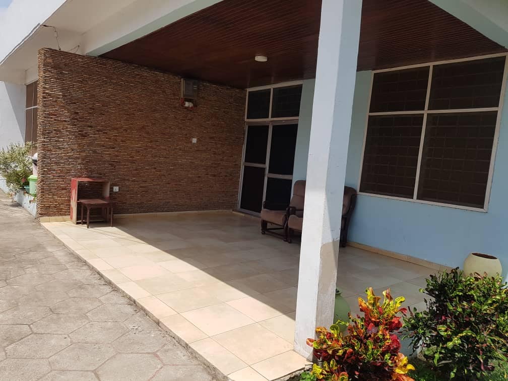 An outdoor seating area is presented with a tiled floor and a ceiling featuring wooden elements. A pair of comfortable chairs sits in the corner next to a small wooden table, while vibrant plants flourish along the edges, creating a welcoming environment.