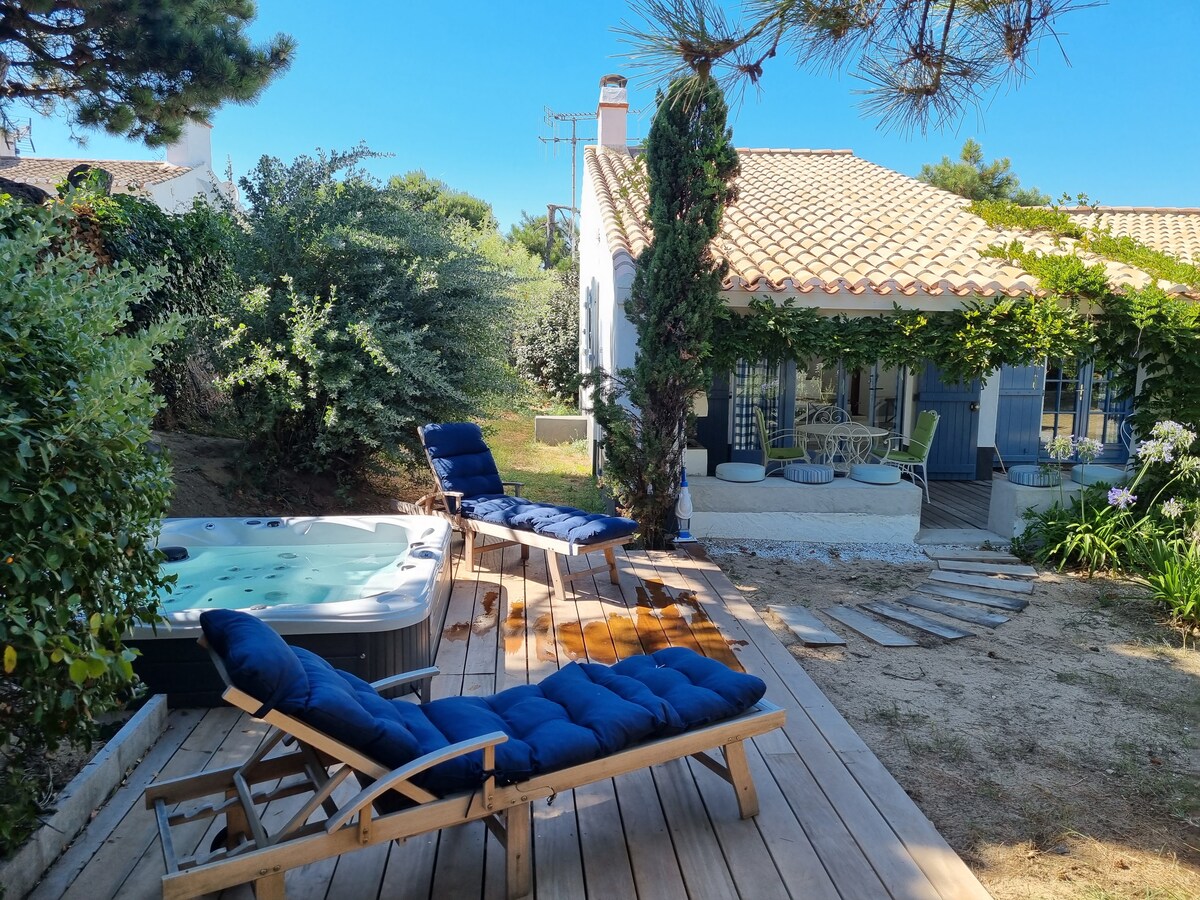 A private jacuzzi is surrounded by wooden decking and paired with two lounge chairs, inviting relaxation. The exterior of the charming white house features a tiled roof and lush greenery, providing a serene atmosphere. A path leads from the jacuzzi area to the entrance.