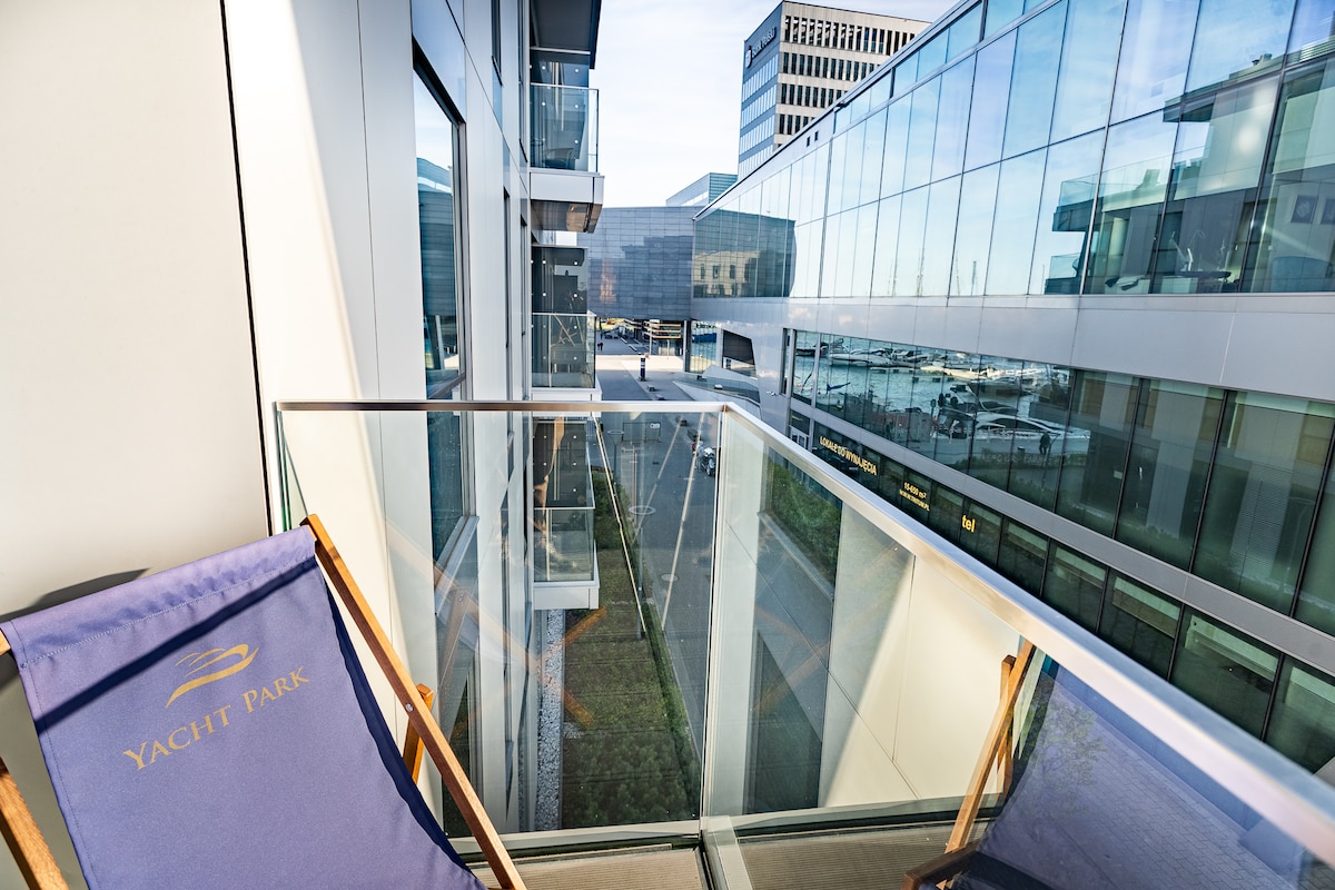 A balcony view is presented, featuring a clear glass railing and a wooden chair adorned with the 'Yacht Park' logo. The surroundings include modern buildings and a glimpse of the marina, enhancing the sense of the urban environment near the waterfront.