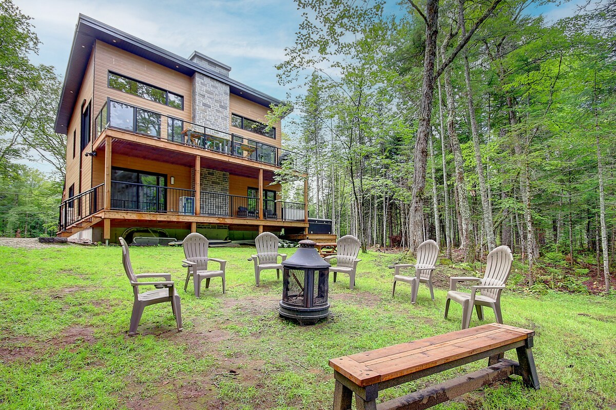 A modern chalet with a wooden deck surrounded by lush trees is visible. Multiple light-colored chairs are arranged in a circle around a central fire pit, complemented by a wooden bench nearby. The structure features large windows, inviting natural light into the space.