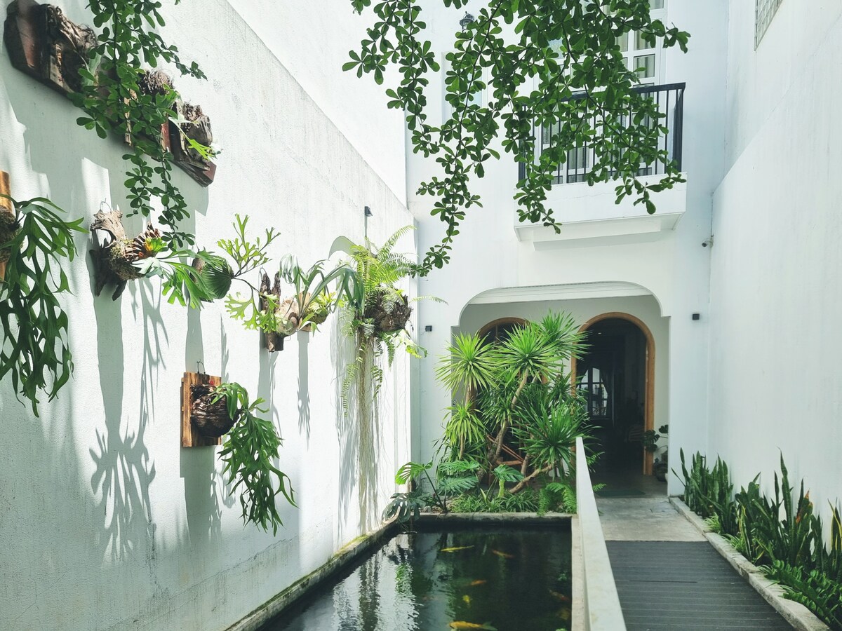 A serene pathway features a well-maintained garden with lush greenery and plants mounted on the walls. A tranquil water feature reflects the surroundings, while a wooden bridge leads to an arched entrance, framed by natural elements and soft lighting.