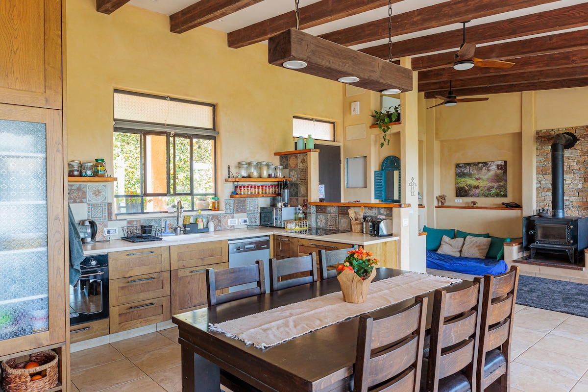 A spacious kitchen is equipped with modern appliances and ample cabinetry. A large dining table with wooden chairs is positioned centrally. Natural light enters through multiple windows, illuminating the warm, earthy tones of the walls and wooden beams overhead.