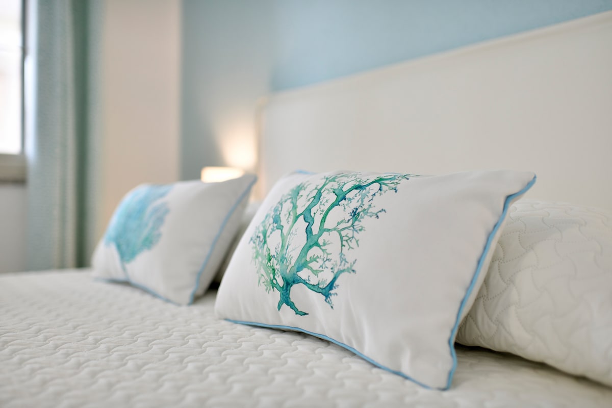 The close-up view of a spacious bed features two decorative pillows with a nature-inspired design. A subtle blue color palette is present on the walls, complemented by soft natural light entering through a nearby window.