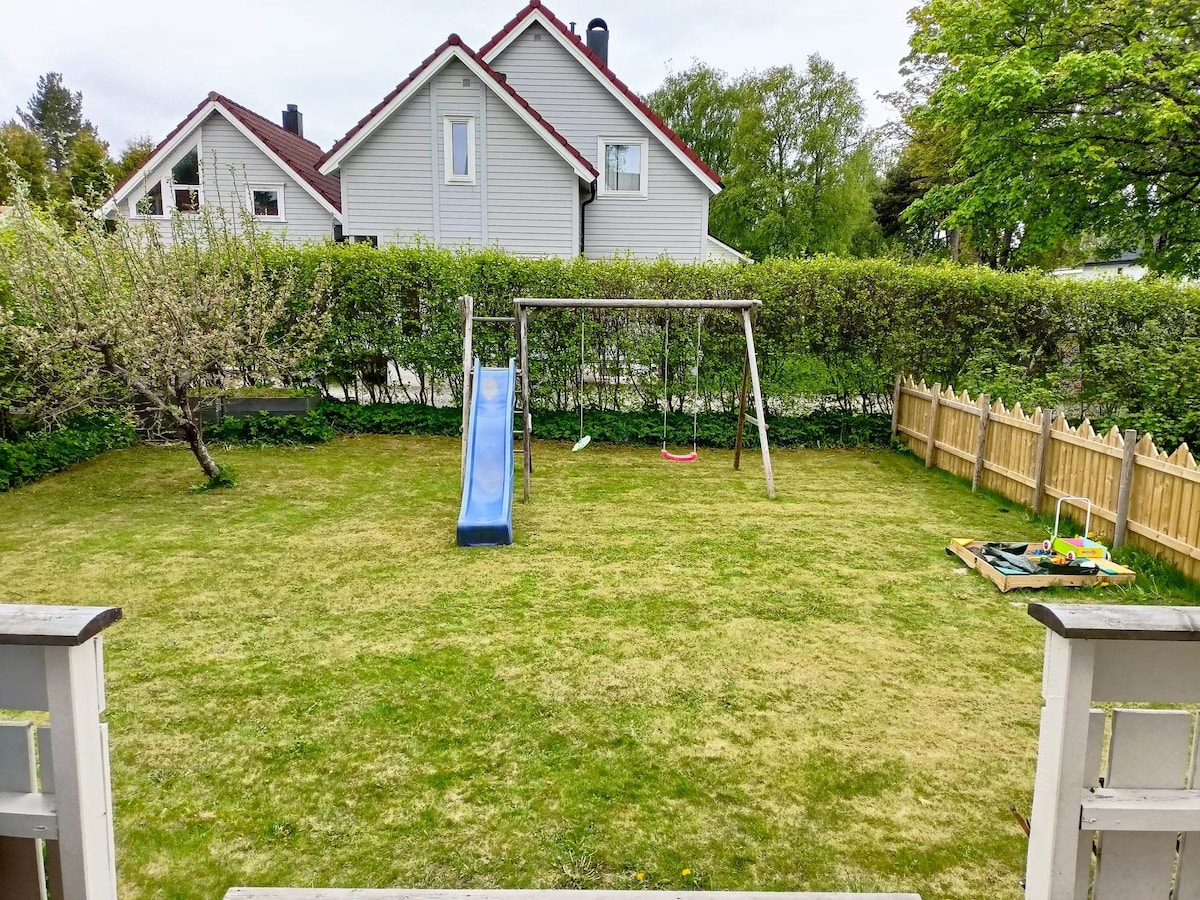 The spacious backyard features a slide and swing set, set against a backdrop of greenery and a wooden fence. A small play area with toys is located to the side, while a well-maintained lawn provides open space for outdoor activities.