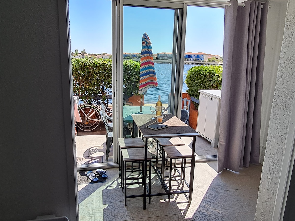 A modern dining area features a small table accompanied by two stools, positioned near large glass doors that open to a lakeside view. A colorful umbrella rests in the center of the table, with a bottle of wine and a notebook nearby, providing a calming atmosphere.