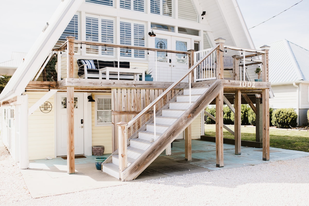 The exterior of a charming A-frame house is shown, featuring a raised deck with seating. Stairs lead to the entrance, while bright white railings enhance the welcoming appearance. Surrounding greenery and a light gravel area offer a tranquil setting.