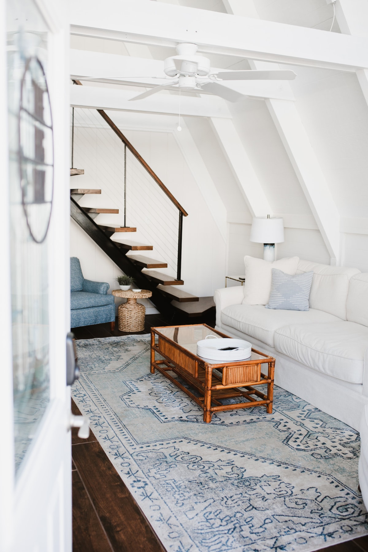 An inviting living area is highlighted by a light-colored sofa and a wooden coffee table resting on a patterned area rug. A ceiling fan and natural light from the entrance enhance the bright, airy atmosphere. A staircase leads to the upper level.