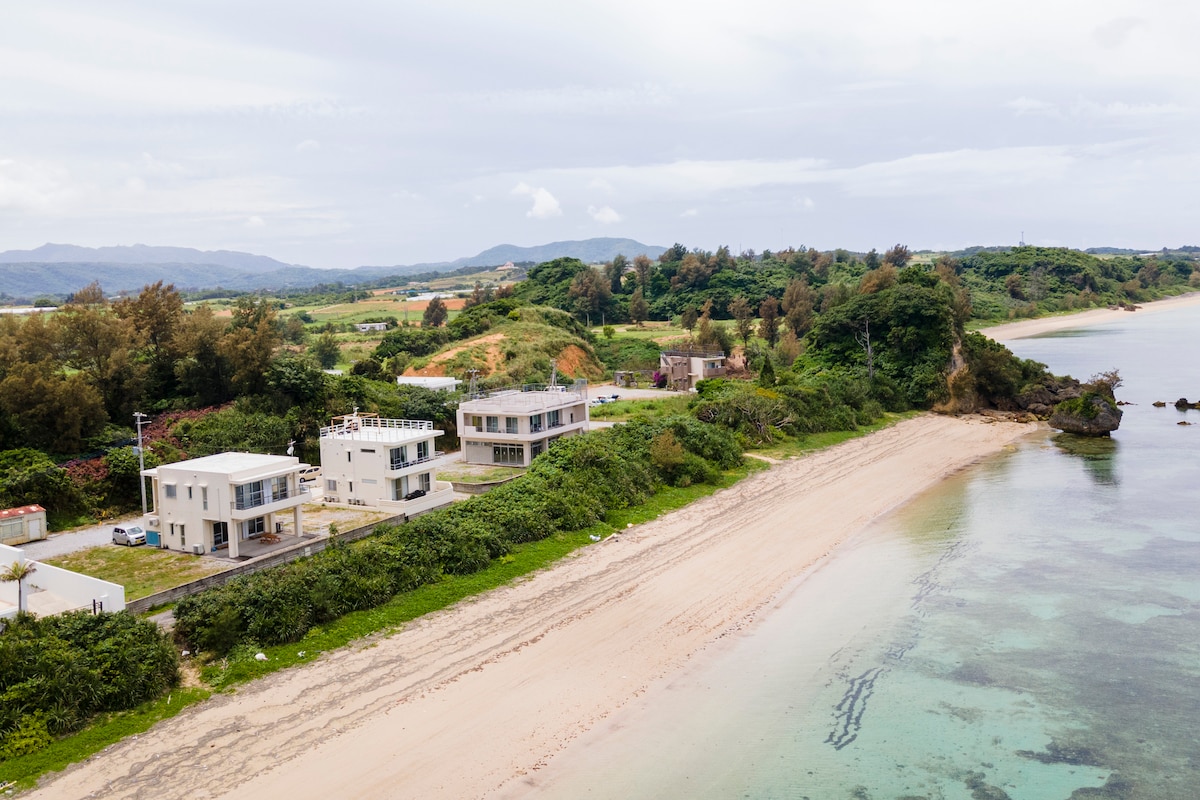 A serene view of the natural beach is captured, with a coastline marked by soft sand and gentle waves. Nearby, a few buildings stand amidst lush greenery, with the backdrop of rolling hills and a cloudy sky, creating a peaceful environment.