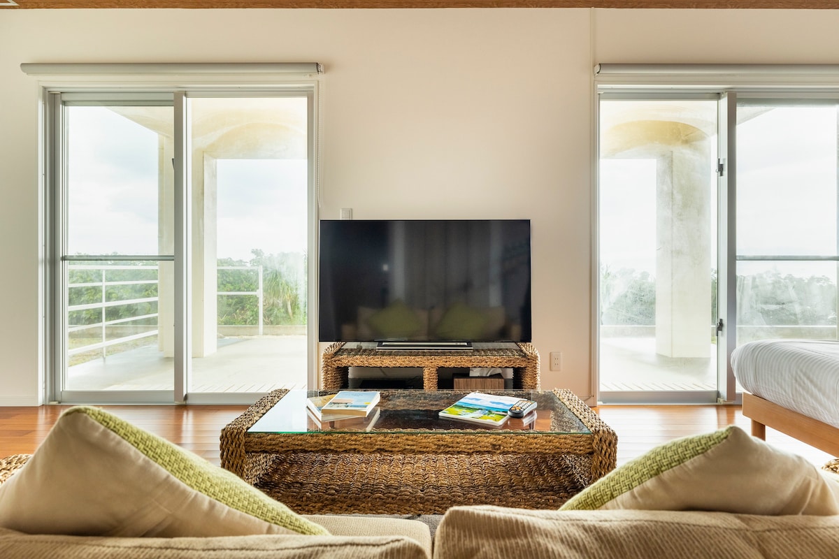 A living area is presented with a large TV and a low, woven coffee table. Two soft seating areas are arranged invitingly. Large glass doors offer views of the outdoor balcony, allowing natural light to fill the space.