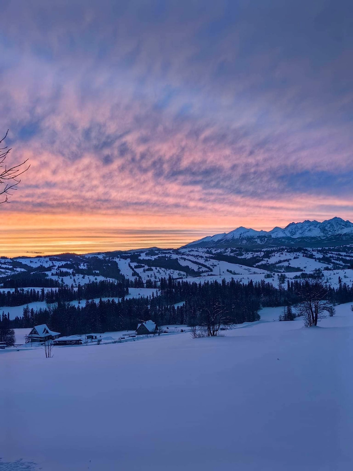 A winter landscape showcases a vast expanse of snow-covered hills, framed by dark evergreen trees. The sky is illuminated with soft hues of pink and purple, transitioning to blue, as the sun sets behind prominent mountain peaks. The scene evokes a serene and peaceful atmosphere.