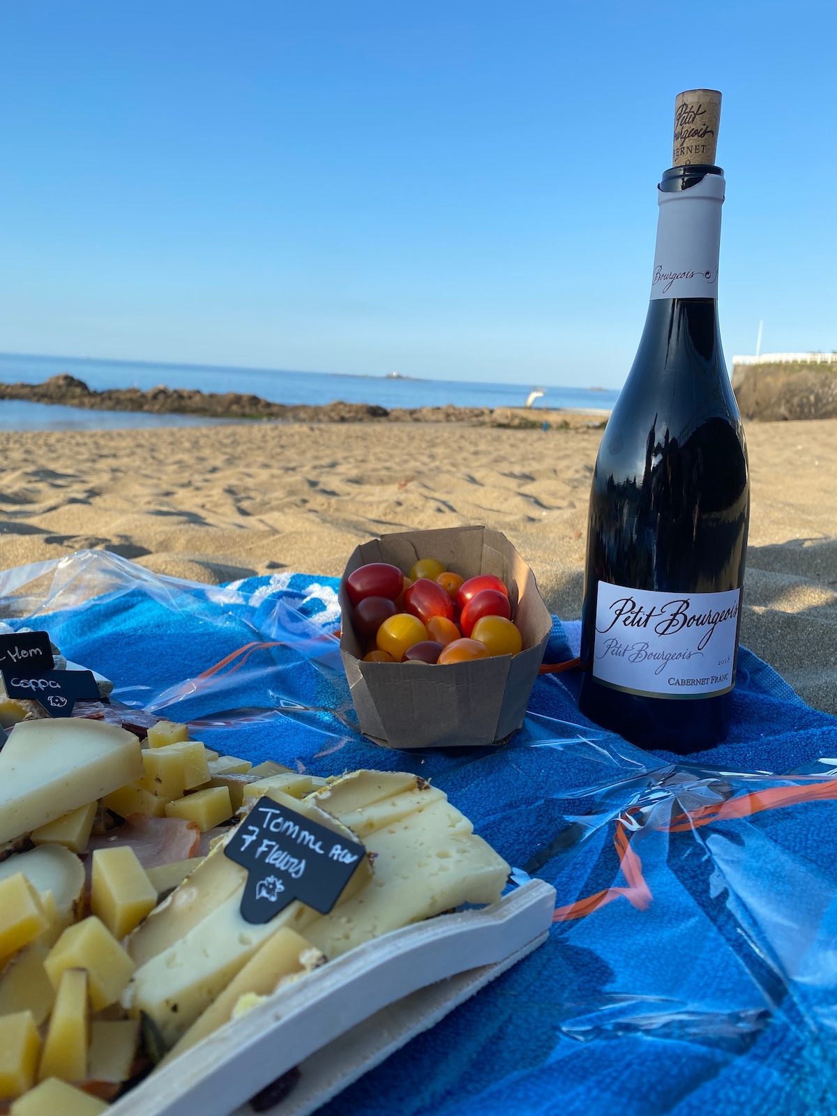 A beach scene captures a spread of assorted cheeses and colorful tomatoes arranged on a blue blanket. A bottle of wine stands nearby, with the ocean visible in the background under clear skies.