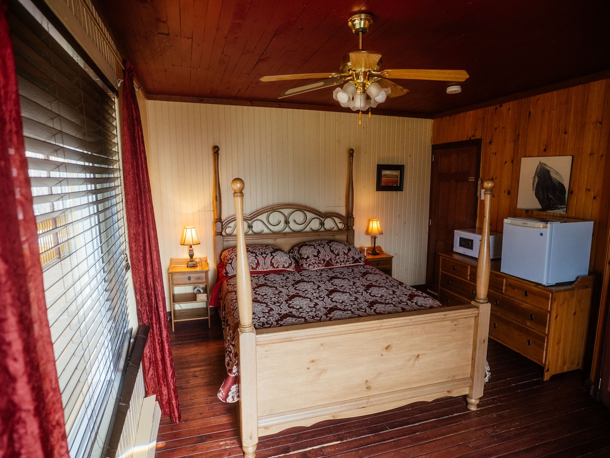 A comfortable bedroom is featured with a wooden bedframe and a floral-patterned bedspread. Two bedside lamps provide soft lighting. A dresser and a microwave are visible, along with a ceiling fan above. Natural light enters through the window covered by vertical blinds.