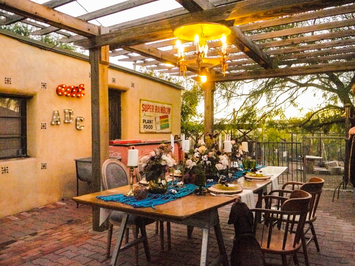 An outdoor dining area is featured under a wooden pergola, with a long table set for a meal. Decorative candles and flowers adorn the table, while soft lighting from a chandelier enhances the ambiance. A view of the surrounding landscape is visible in the background.