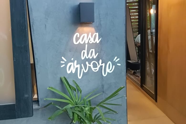 The exterior wall of the Casa da Árvore displays the name 'casa da árvore' in a playful script. A wall-mounted light provides illumination, while a green potted plant is positioned at the base, adding a touch of nature to the entrance area.
