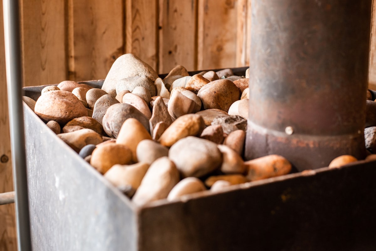 A close-up view showcases a wood-fired sauna stove, surrounded by an array of smooth, river stones. The stones vary in size and color, resting inside a sturdy, metal base connected to a chimney. Natural wooden walls create a rustic ambiance.