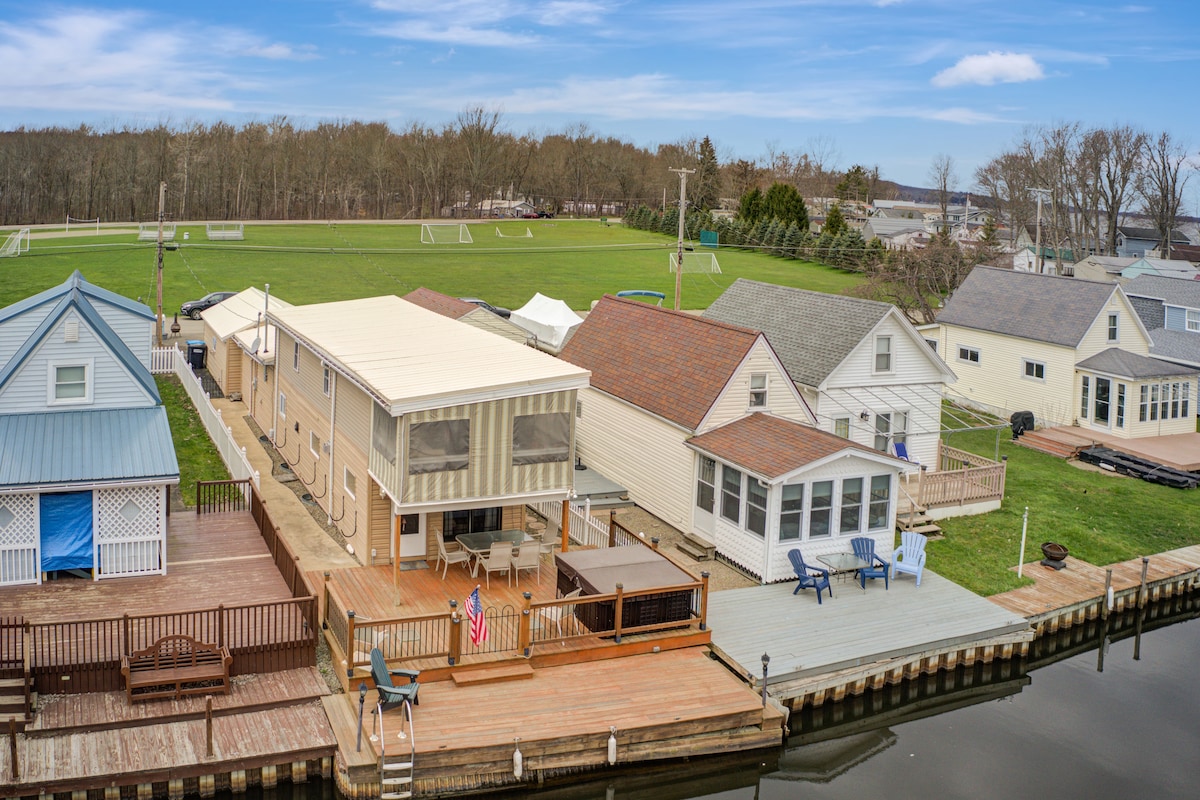 An aerial view of a waterfront cottage reveals a spacious deck with a hot tub, seating areas, and a private dock along the canal. Adjacent green fields and nearby structures contribute to the inviting outdoor environment.