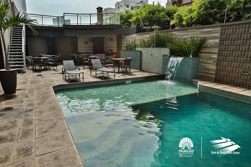 A clear swimming pool is surrounded by a tiled patio featuring lounge chairs and tables. A gently flowing water feature adds a tranquil sound, while greenery provides natural accents around the space. Stairs lead up to an upper level, enhancing accessibility.