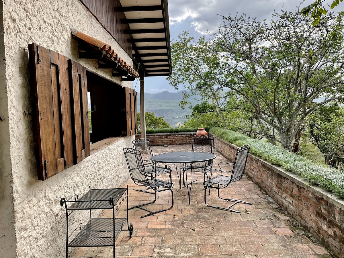 An outdoor terrace is detailed with a round metal table surrounded by several chairs. Stone walls and wooden shutters frame the view, where greenery and distant mountains are visible. The atmosphere is tranquil, perfect for relaxation or social gatherings.