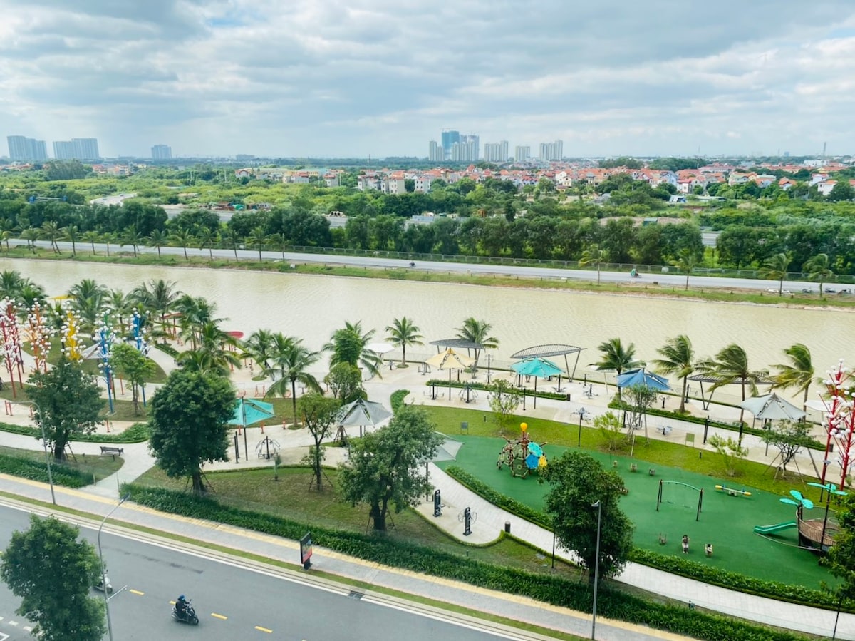 The image presents a view of a landscaped riverfront park, featuring pathways, palm trees, and play areas. The river flows calmly beside the park, with greenery extending into the distance. A mix of modern buildings is visible on the horizon under a partly cloudy sky.