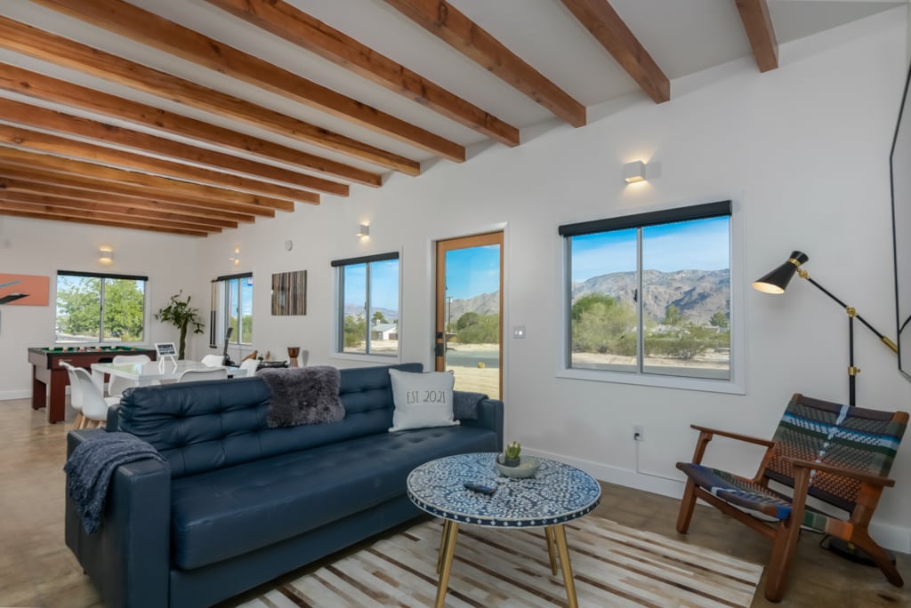 The living area is characterized by vaulted wooden ceilings and large windows that provide natural light and mountain views. A dark sofa is complemented by a patterned area rug and a round coffee table, while a dining area is visible in the background.