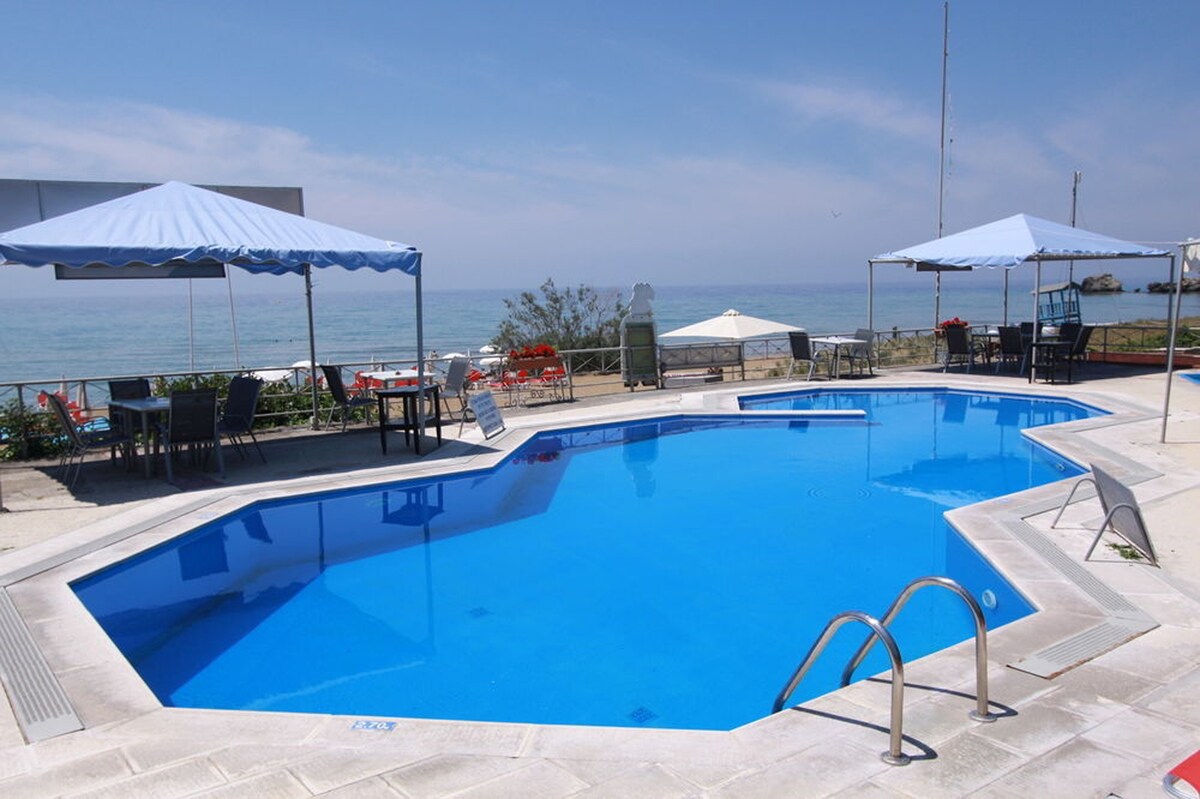 A spacious outdoor pool is surrounded by sun loungers and umbrellas, providing shade and relaxation. The crystal-clear water reflects the blue sky, while the beach can be seen in the background, enhancing the tranquil atmosphere of the area.