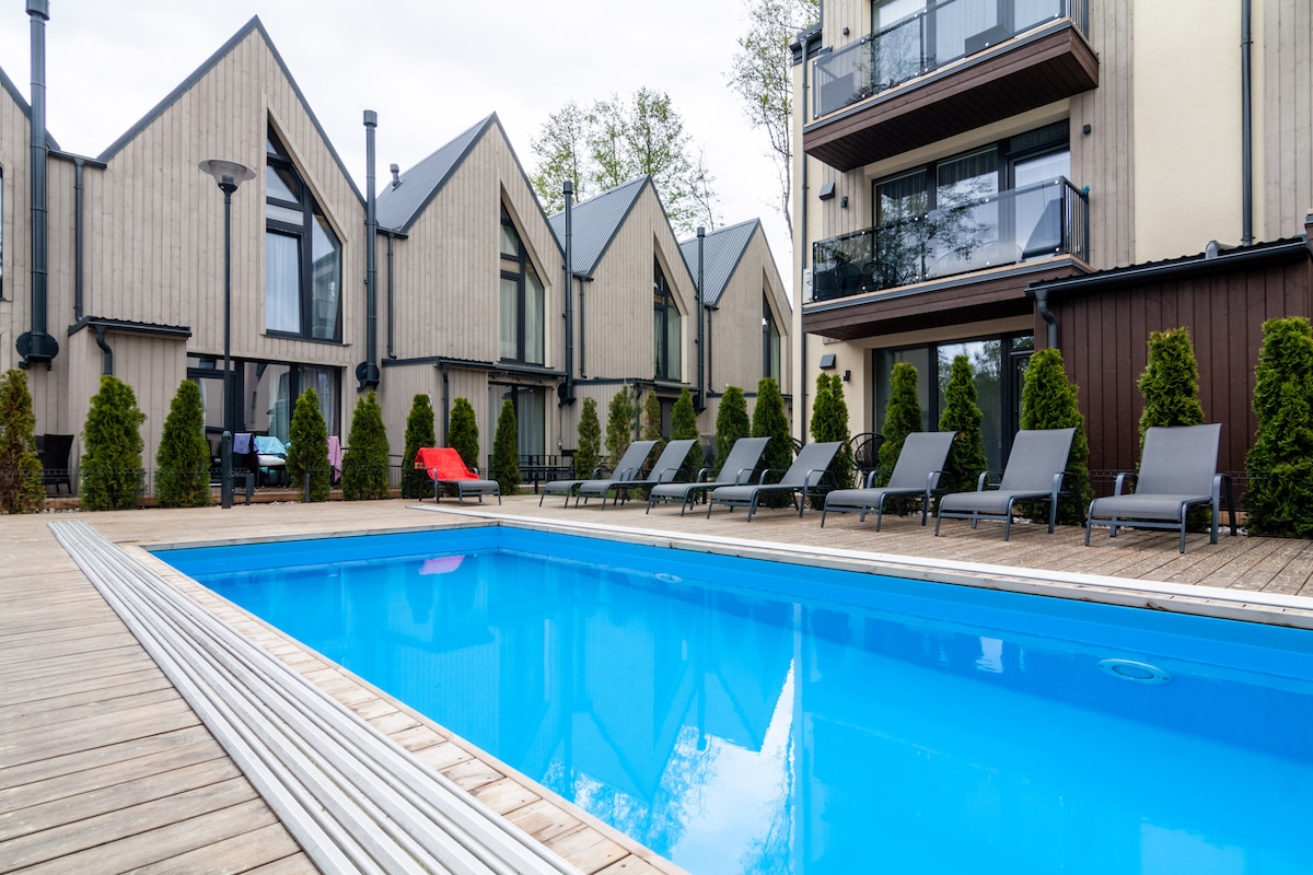 A clear blue swimming pool is surrounded by a wooden deck, with multiple lounge chairs positioned on one side. The area features a backdrop of modern buildings with balconies and manicured greenery, creating a serene outdoor setting.