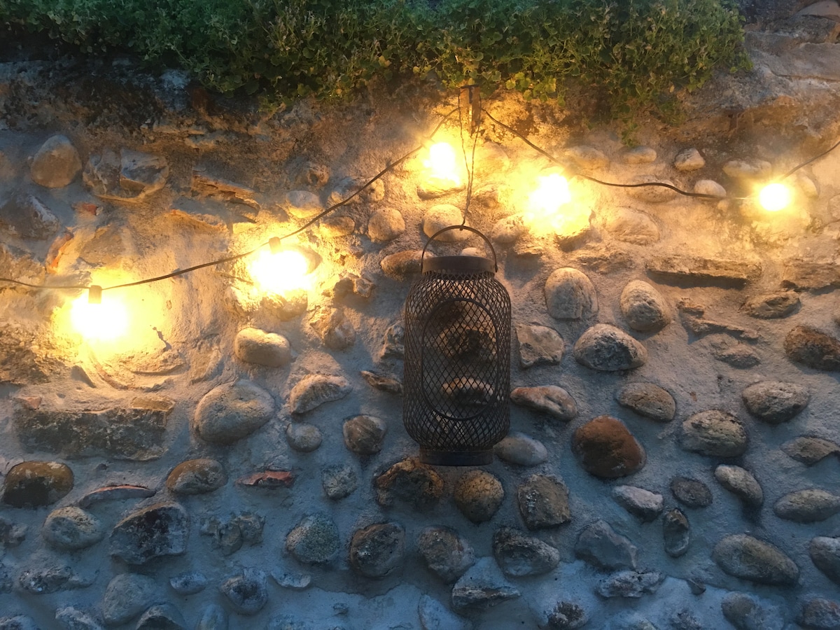A rustic stone wall is adorned with warm glowing string lights and a metal lantern, creating a cozy ambiance. The textures of the stones are highlighted, showcasing a natural and inviting outdoor space.