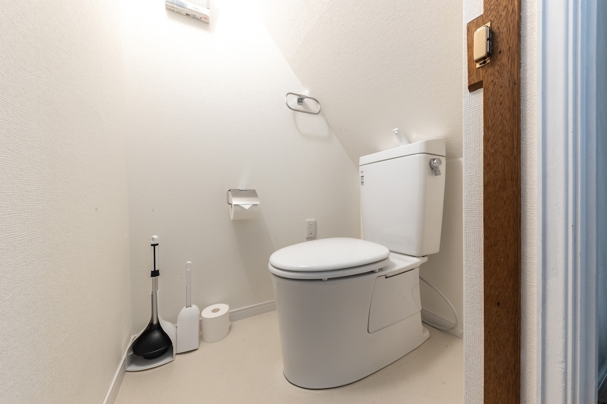 A clean and functional bathroom is shown, featuring a modern toilet with a sleek design. Adjacent to the toilet, a toilet brush and plunger are placed for convenience. Soft lighting enhances the bright, neutral wall color, creating a spacious feel.