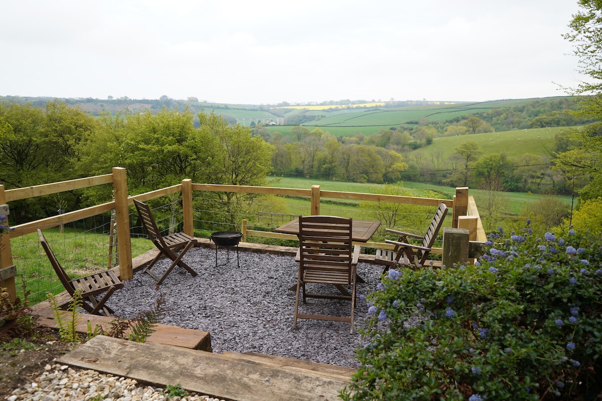 An outdoor seating area is presented with wooden furniture, including a table and chairs, set on a gravel surface. Surrounding greenery and distant hills create a scenic backdrop, while a wooden railing offers a clear view of the countryside.