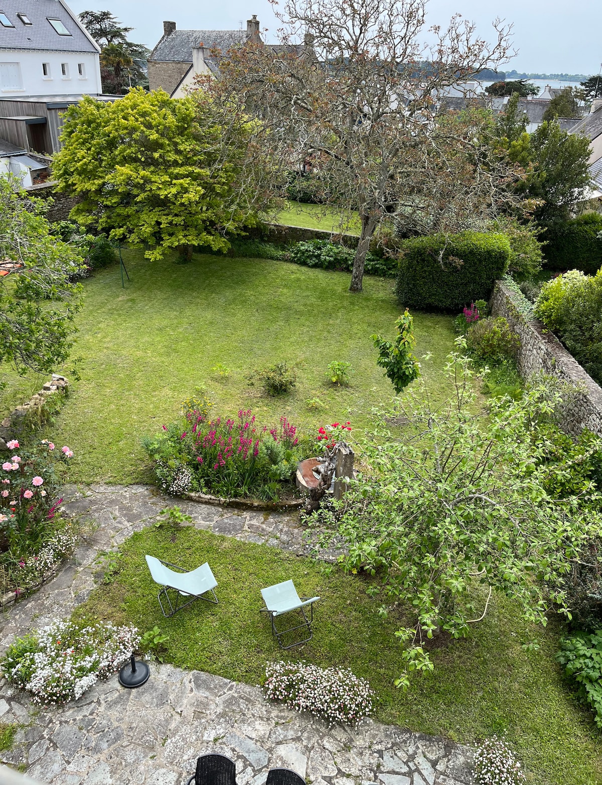 A landscaped garden is viewed from above, featuring a lush green lawn surrounded by colorful flowerbeds. Two light blue chairs are positioned on a stone patio, inviting relaxation. Mature trees and various plants add to the garden's natural ambiance.