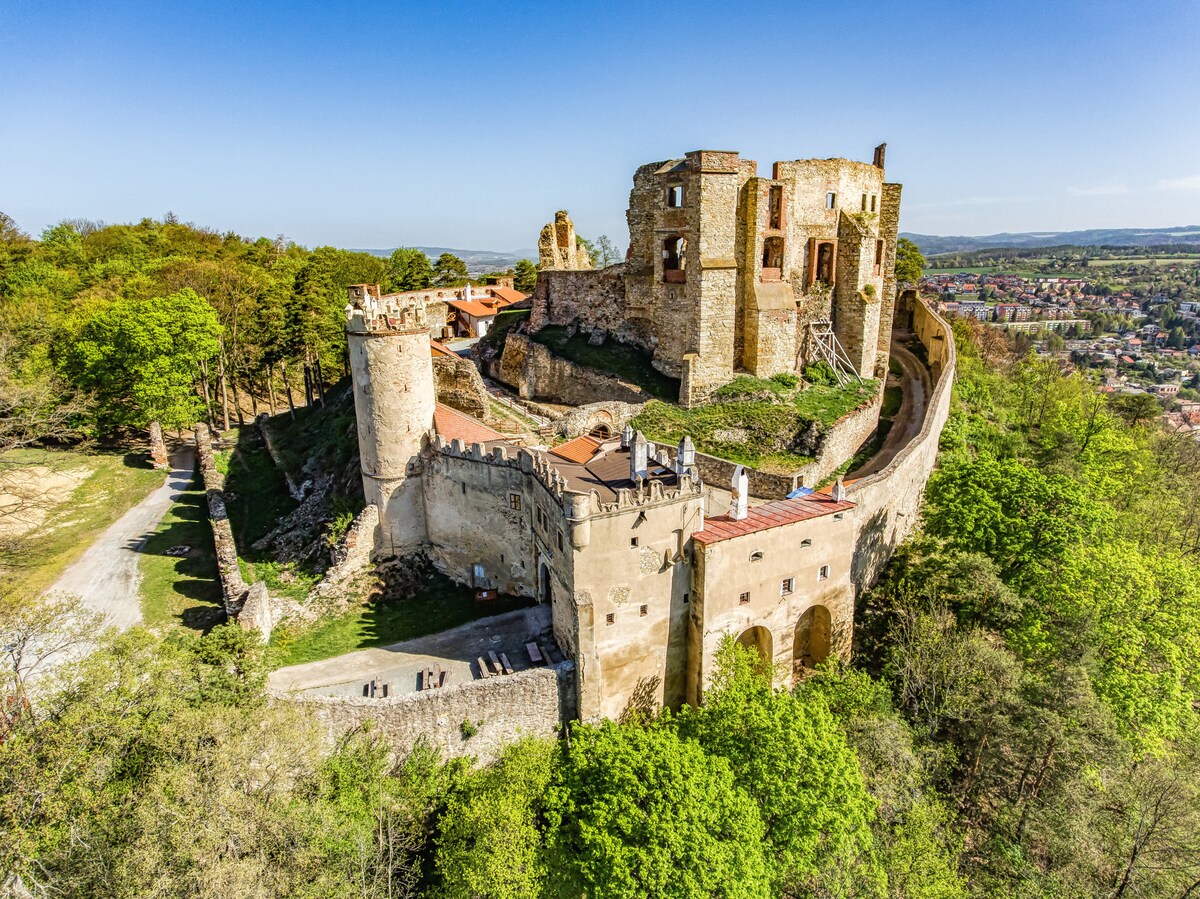 Aerial view of a historical castle ruin situated on a hilltop, surrounded by lush greenery. The stone structure features multiple towers and walls, showcasing the remnants of its former grandeur. A clear blue sky frames the scene, emphasizing the natural setting.