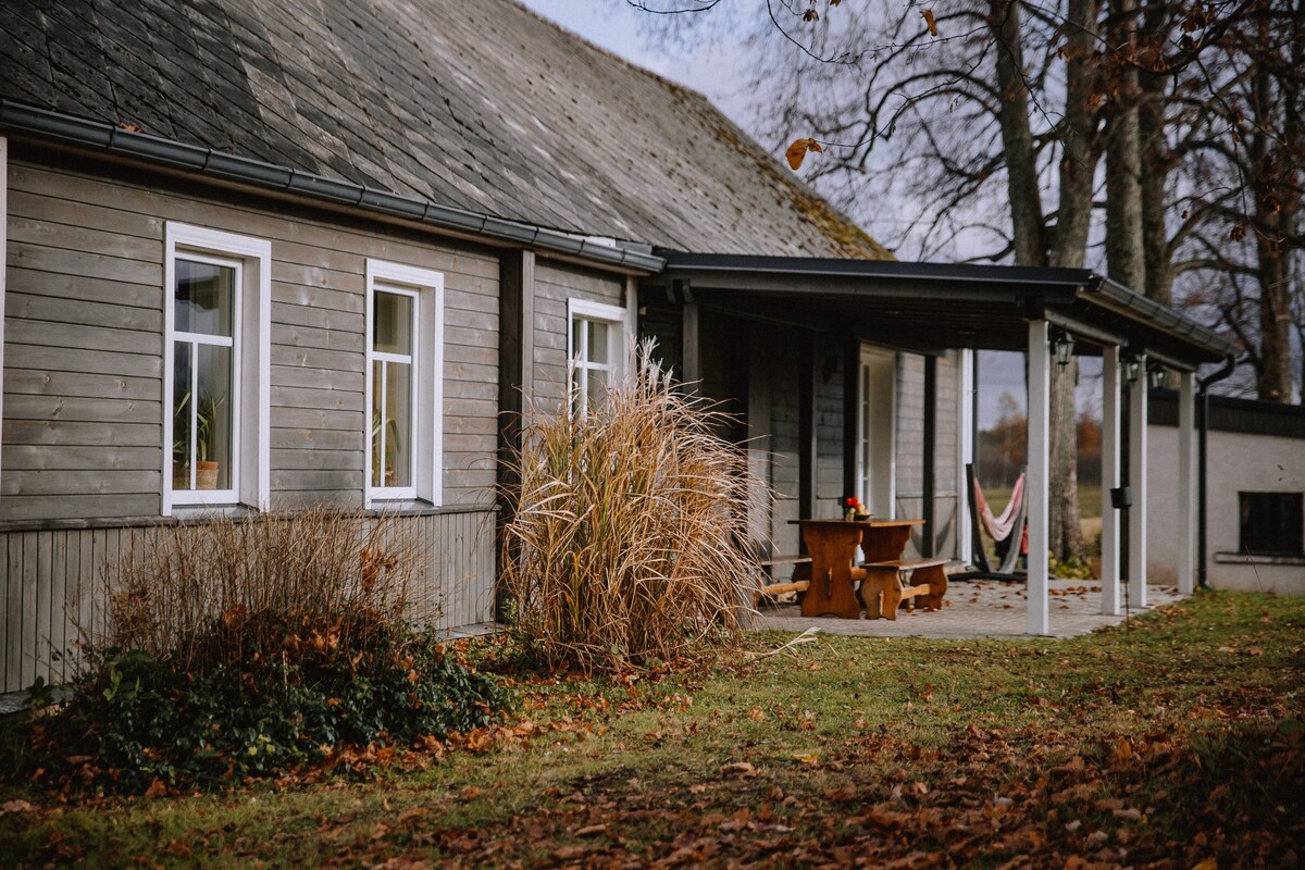 The exterior of a restored rural home is presented, featuring wooden siding and a sloped roof. A shaded porch is visible with a wooden table and seating. Lush grass and autumn foliage surround the structure, enhancing the serene setting.