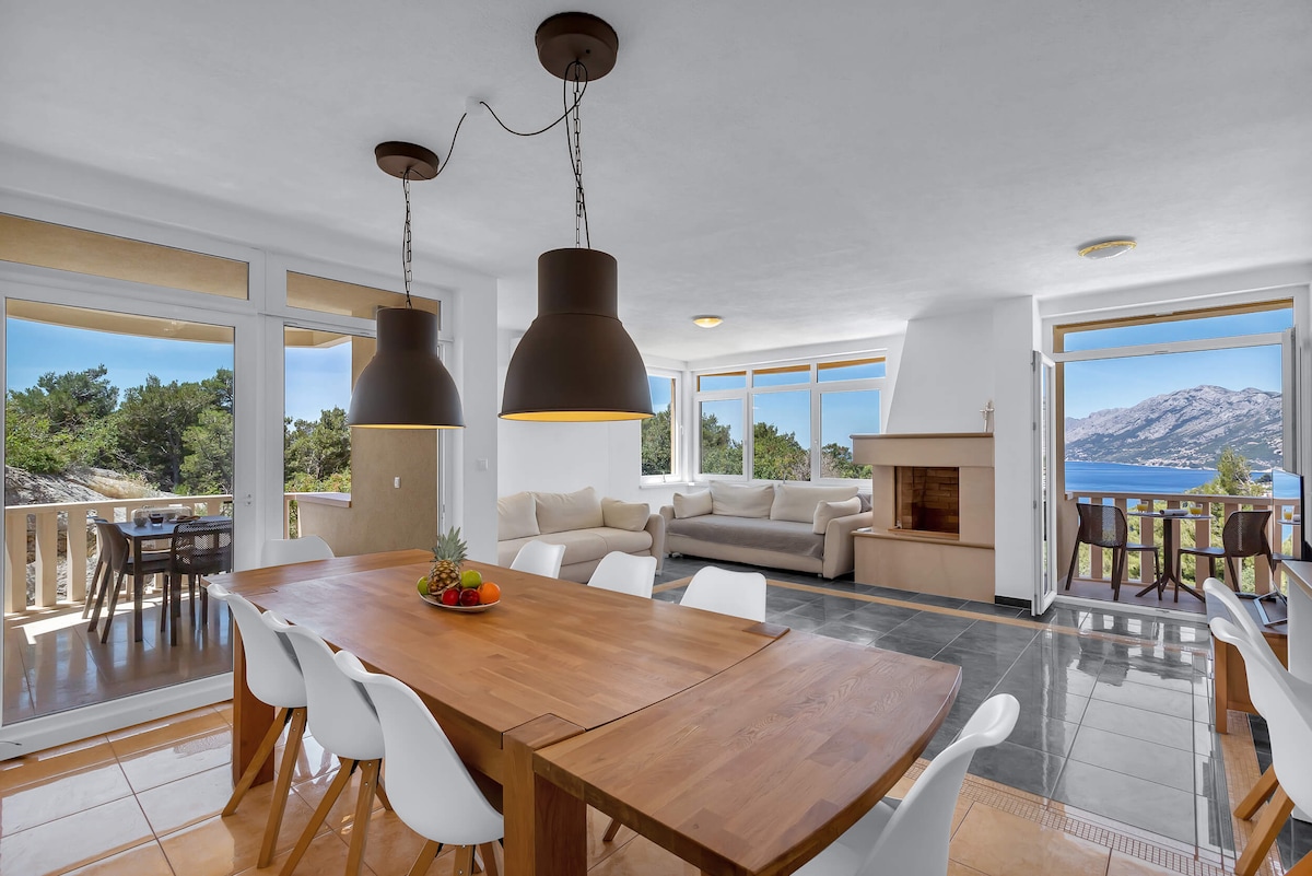 A spacious living area features large windows providing views of the surrounding landscape. A dining table with white chairs sits prominently, accompanied by a cozy seating area. An indoor fireplace is visible, contributing to the room's function as a gathering space.