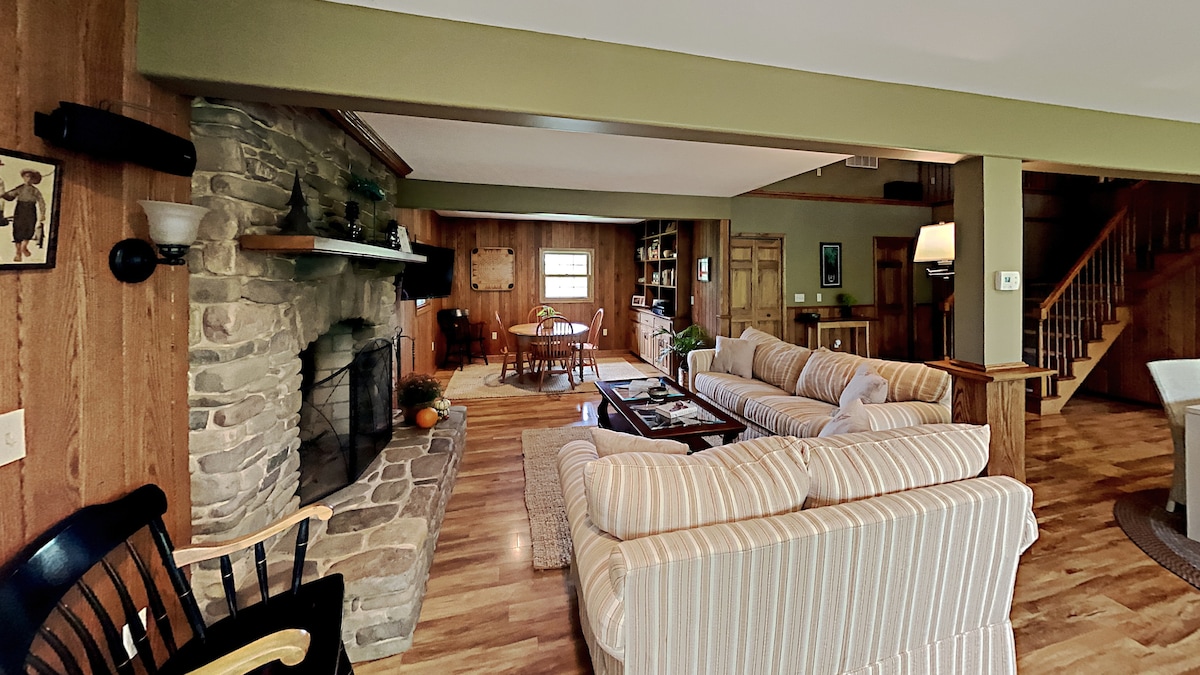 A spacious living area features a stone fireplace, surrounded by comfortable seating. Natural light enters through a large window, highlighting wooden paneling and flooring. A dining area is visible in the background, accompanied by a bookshelf, creating a welcoming environment for relaxation and gatherings.
