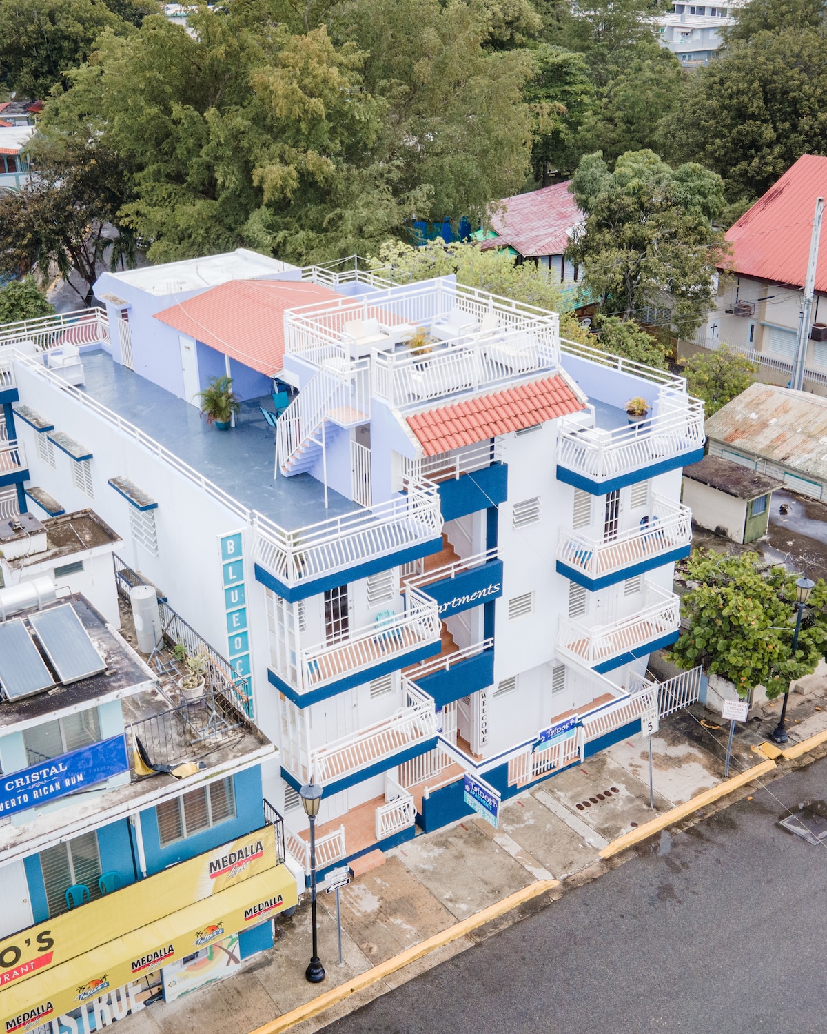 An aerial view of a three-story building showcases its modern architecture, featuring blue and white exteriors. Multiple balconies with railings and potted plants are visible, along with a staircase leading to the top floor. Surrounding greenery and nearby structures are included in the vibrant urban setting.