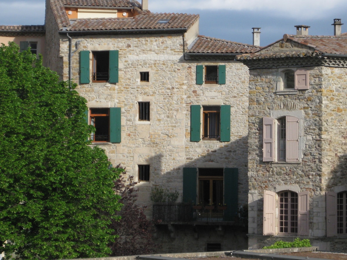 The image showcases a charming stone building featuring multiple windows with green shutters and wooden balconies. The structure is complemented by nearby trees providing greenery, set against a backdrop of a cloudy sky.