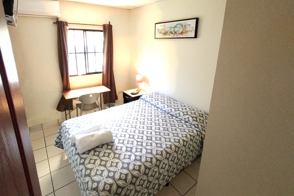 A cozy room is showcased, featuring a double bed with a patterned duvet cover. A desk with a chair is positioned by the window, complemented by a bedside lamp. Natural light enters through the window, while tiled flooring adds to the room's functionality.