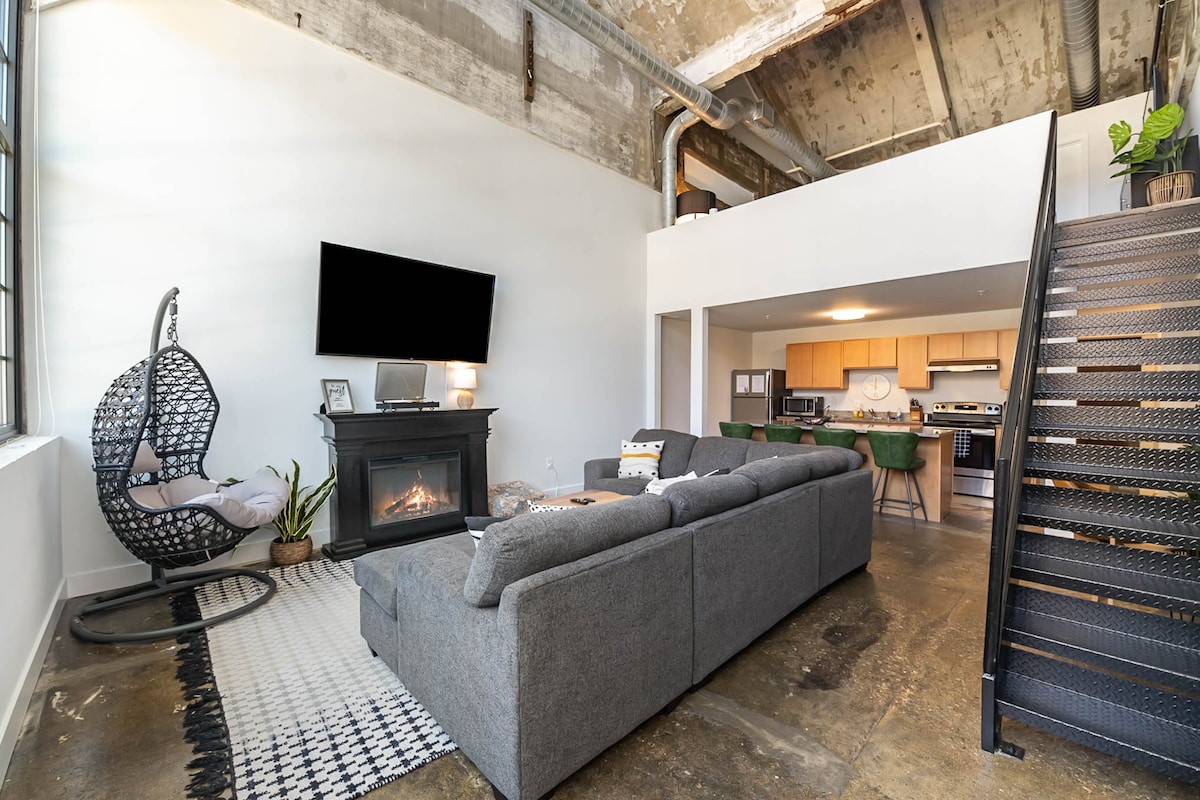 The open living area features a spacious couch facing a modern television and an electric fireplace. A round dining table sits in the background, while a striking black staircase leads to the loft above. Natural light floods in through large windows, highlighting the concrete flooring.