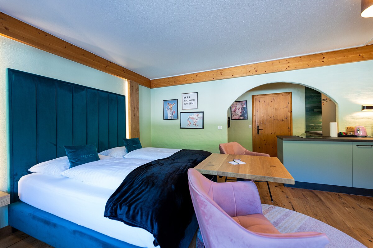 The modern studio apartment features a spacious bed with a dark throw blanket, complemented by a green accent wall. A wooden dining table is paired with pink chairs, creating a cozy dining space. The adjoining kitchen area is subtly visible through an arched opening.