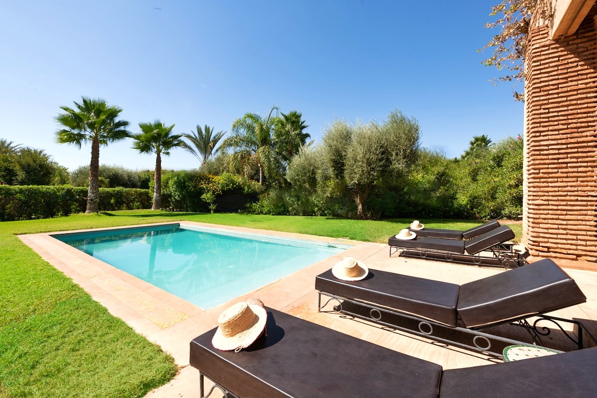 A private pool is set within the lush garden, framed by palm trees and greenery. Two lounge chairs are positioned beside the pool, each adorned with a stylish sun hat. The tranquil atmosphere is enhanced by clear blue skies and sunlight reflecting off the water.