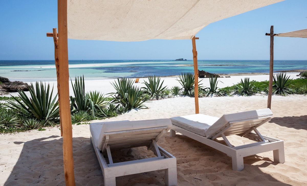 Two white lounge chairs are positioned under a sunshade on the sandy beach, facing the calm turquoise waters. Tropical plants frame the scene, and the shoreline extends into the distance, complemented by a clear blue sky.
